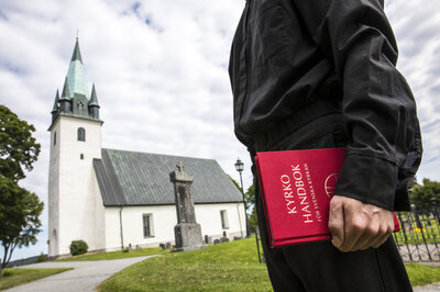 Man i svart skjorta står framför en kyrka med kyrkohandboken i handen.