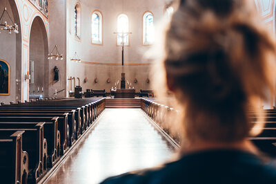 Kvinna står i längst bak i mittgången av en kyrka vänd emot altaret. Solen lyser in bakom Jesus krucifixet över altaret.
