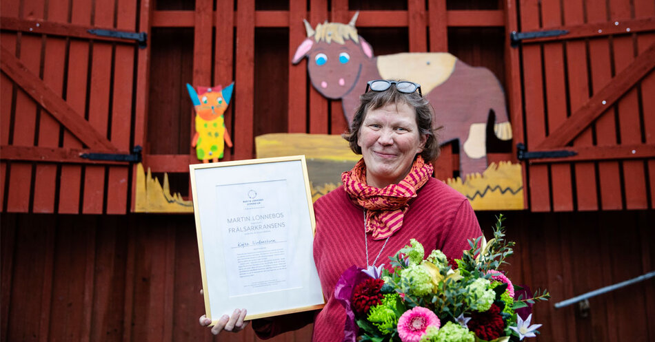 Leende kvinna i röda kläder står framför en röd byggnad och håller ett diplom samt en bukett blommor.