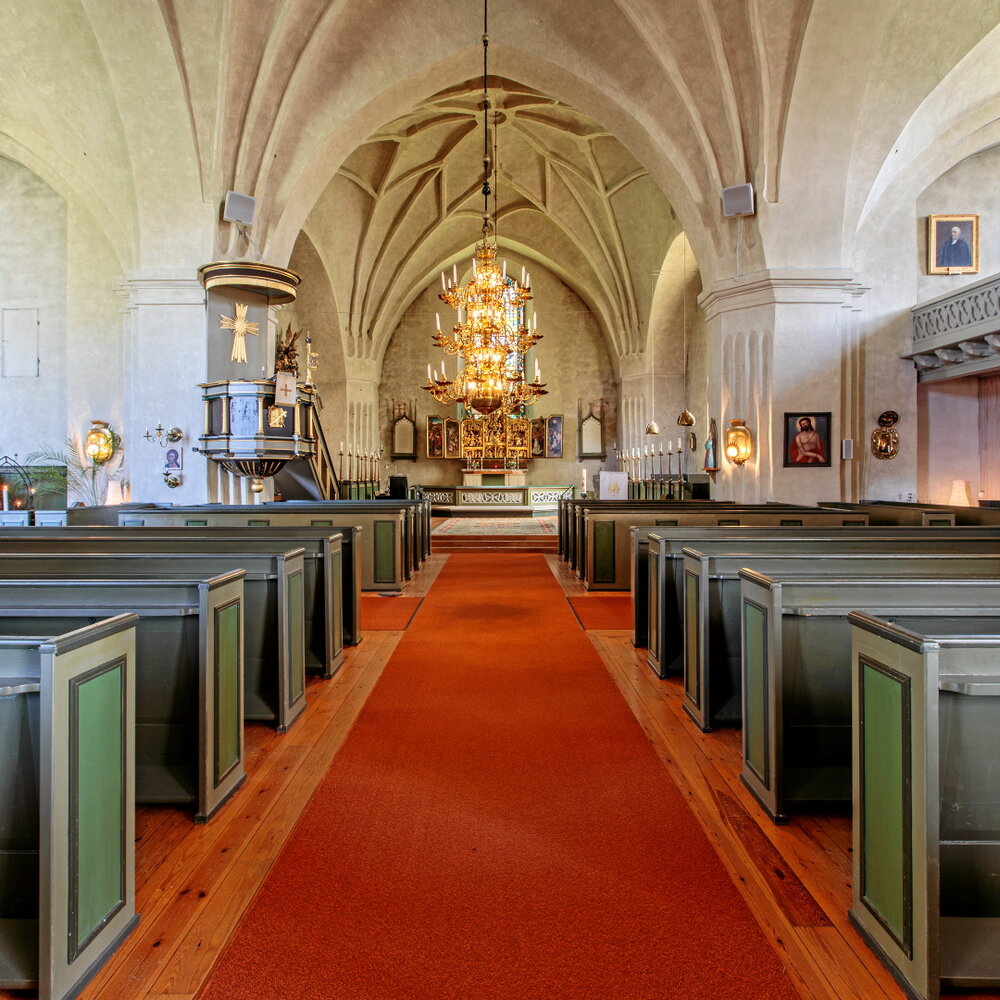 Kyrka fotad inifrån ifrån mittgången. Röd matta, gröna kyrkbänkar samt en stor gyllene ljuskrona hängande från taket.