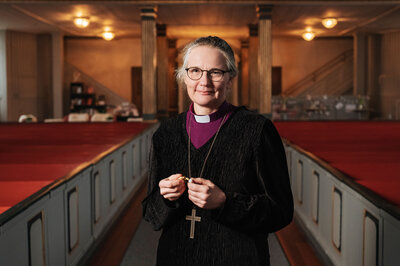 En äldre kvinna i biskopsskjorta och kors står i en kyrka och åller i en Frälsarkrans. | © Foto: Marcus Gustafsson