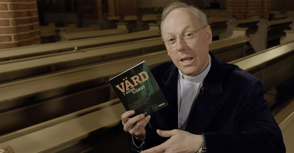 Man i vit prästskjorta sitter i en kyrkbänk och pratar medans han håller upp boken Värd i guds tjänst.