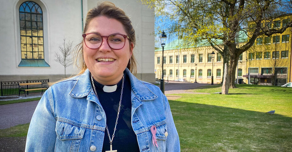 Leende kvinnlig präst i svart prästskjorta med blå jeansjacka över står framför en kyrka. | © Foto: Peter Pasalic Östbort