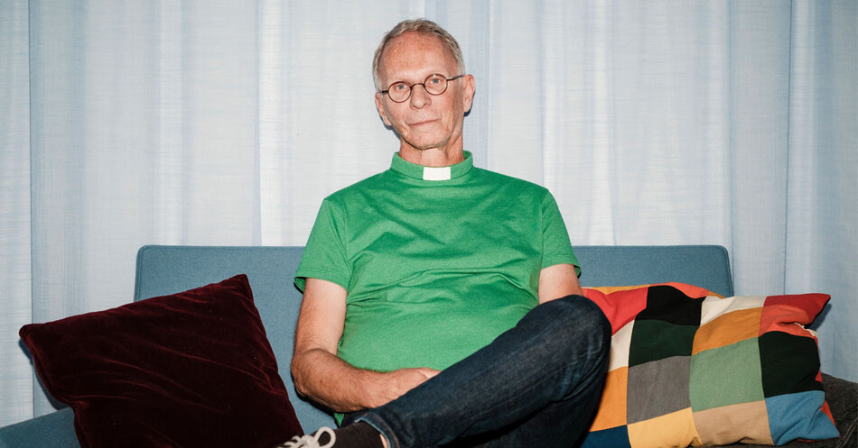Man i grön diakonskjorta med runda glasögon sitter i soffa med benen korsade. | © Foto: Marcus Gustafsson