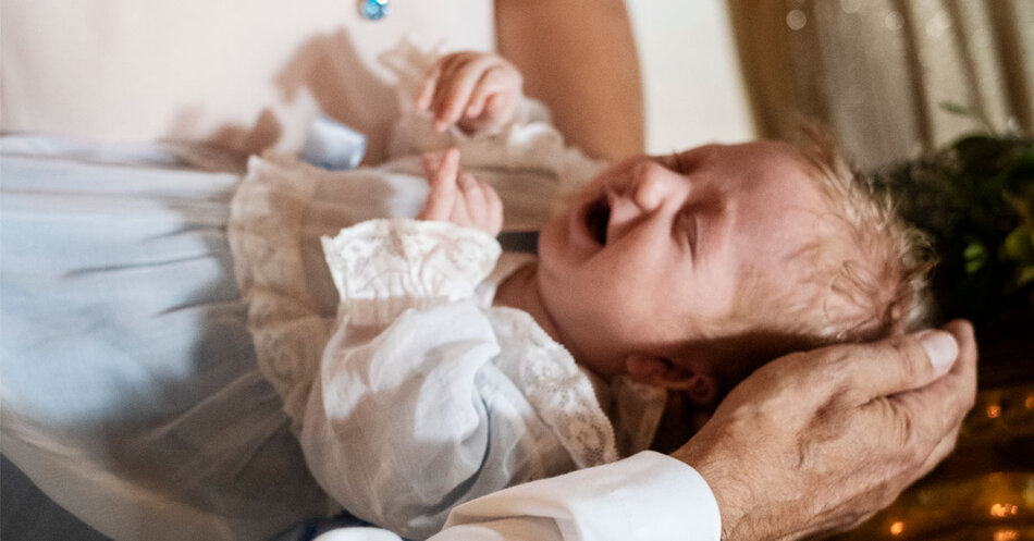 Närbild bebis i ljusblå dopklänning hålls fram till dopfunten medan prästen för vatten till barnets huvud.