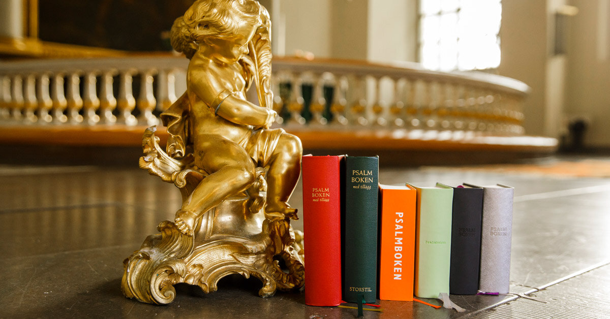 6 olika utgåvor av psalmboken från Verbum uppställda på golvet bredvid en gyllene staty av en bebis på ett kyrkogolv.