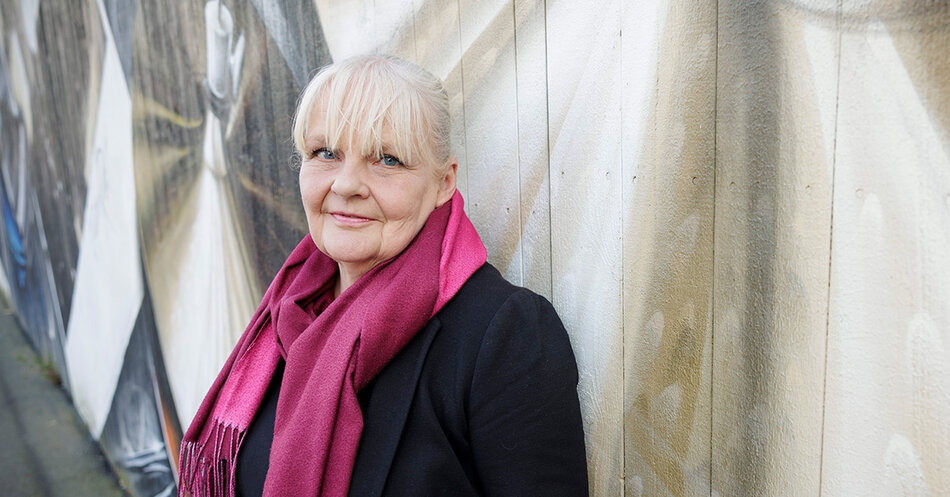 Närbild av en vithårig kvinna med rosa scarf framför en träfasad täckt av ett målat konstverk. | © Foto: Mikael M Johansson