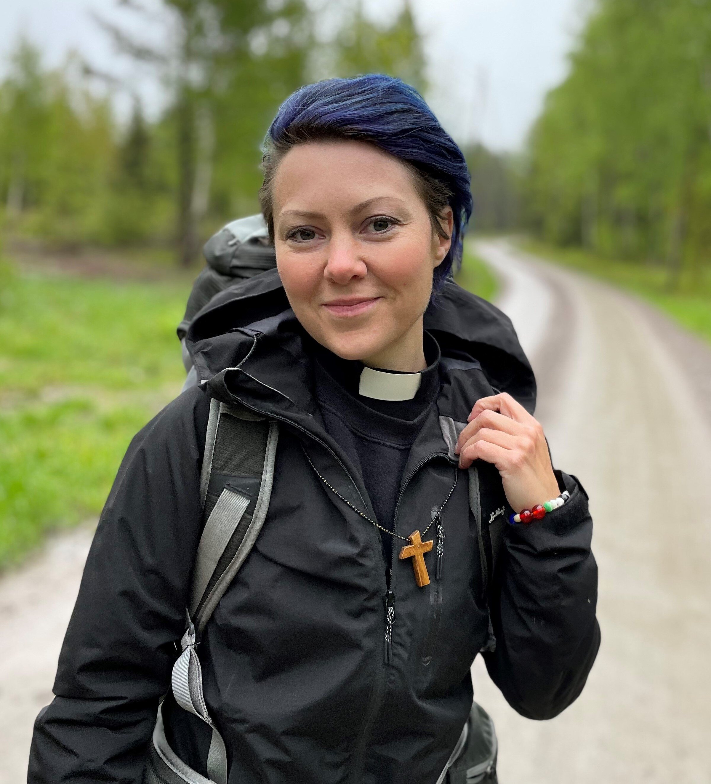 Leende kvinna med blått hår, prästkrage, frälsarkransen och vandringskläder samt ryggsäck står på en grusväg.