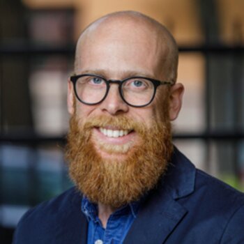 Porträtt närbild av leende man med runda glasögon och stort rött skägg i blå skjorta.