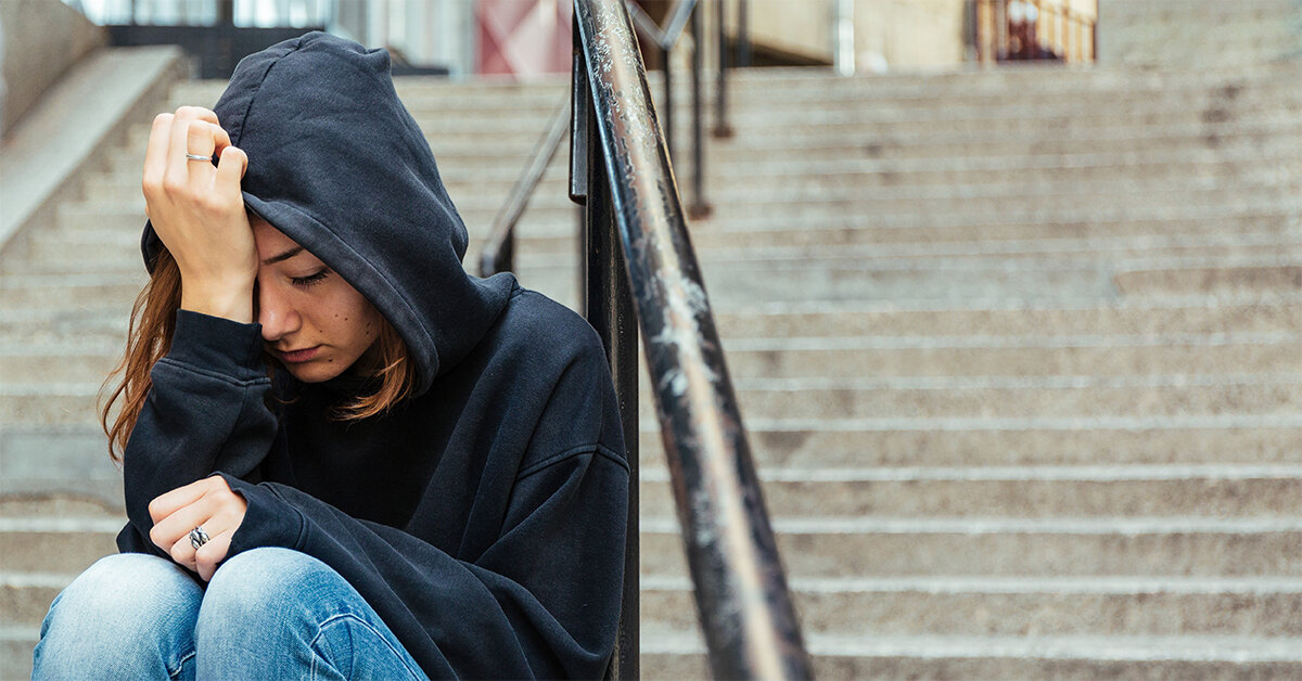 Ungdom iklädd svart hoodie med uppdragen luva sitter med ansiktet lutad emot ena handen i en trappa.