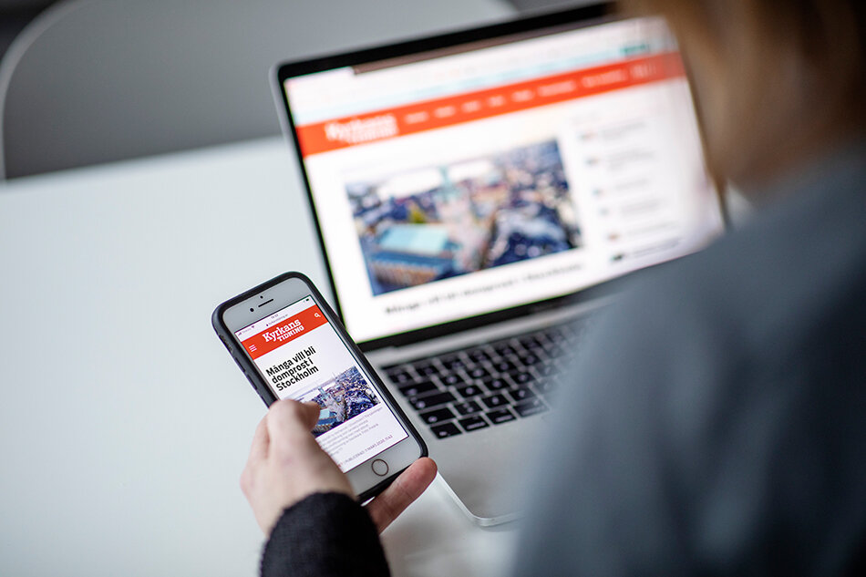 En person sitter framför en laptop med kyrkans tidnings hemsida öppnad och håller en telefon som också visar kyrkans tidnings hemsida 